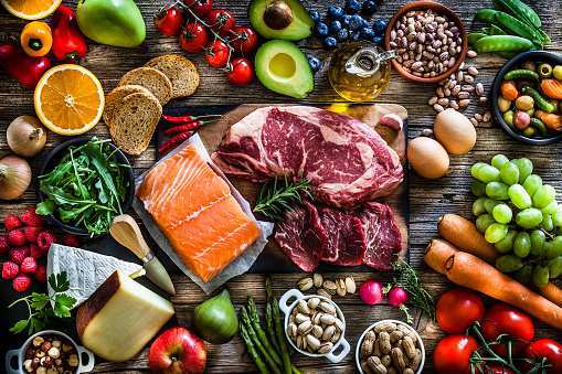 At the center of the frame is a cutting board with beef steak and a salmon fillet and all around it is a large variety of food like fruits, vegetables, cheese, bread, eggs, legumes, olive oil and nuts