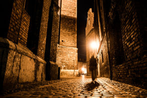 Illuminated cobbled street with light reflections on cobblestones in old historical city by night. Dark blurred silhouette of person evokes Jack the Ripper.